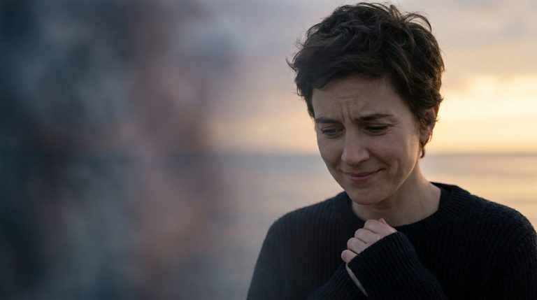 Portrait d'une femme aux cheveux courts, les mains jointes et le regard baissé, semblant pensive ou inquiète devant un coucher de soleil flou.
