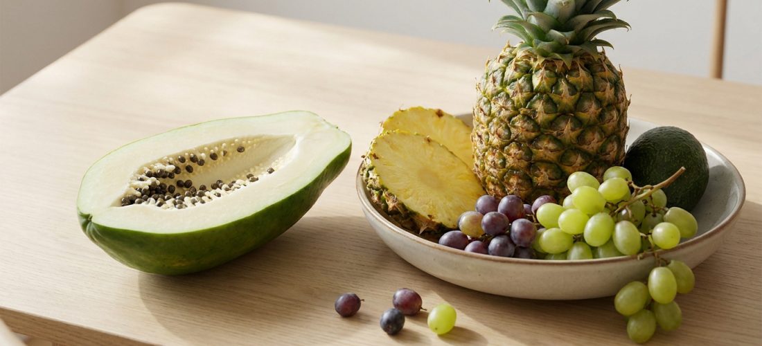 Une papaye verte coupée en deux, un ananas entier et des tranches, des raisins et un avocat sur une table en bois clair.
