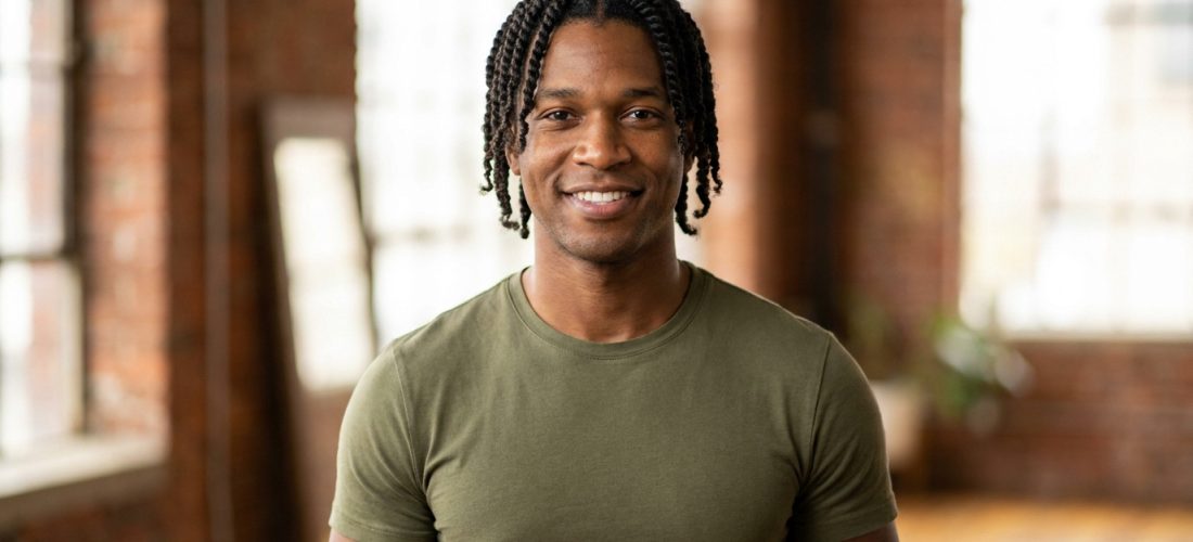 Jeune homme noir souriant portant une coiffure en vanilles, posant dans un loft lumineux aux murs en briques.