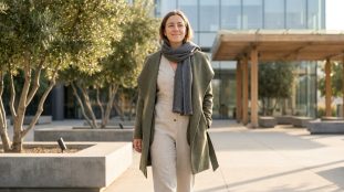 Femme souriante en manteau kaki, combinaison beige et écharpe grise, marchant dans un jardin urbain moderne aux arbres d'oliviers.