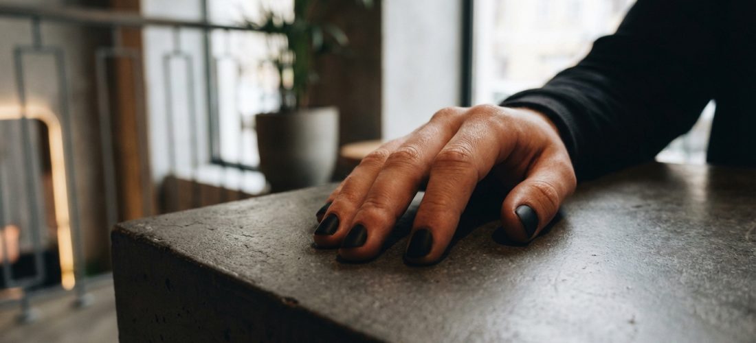 Main d'un homme posée sur une surface sombre, ses ongles sont peints d'un vernis noir mat. Arrière-plan flou, lumineux.