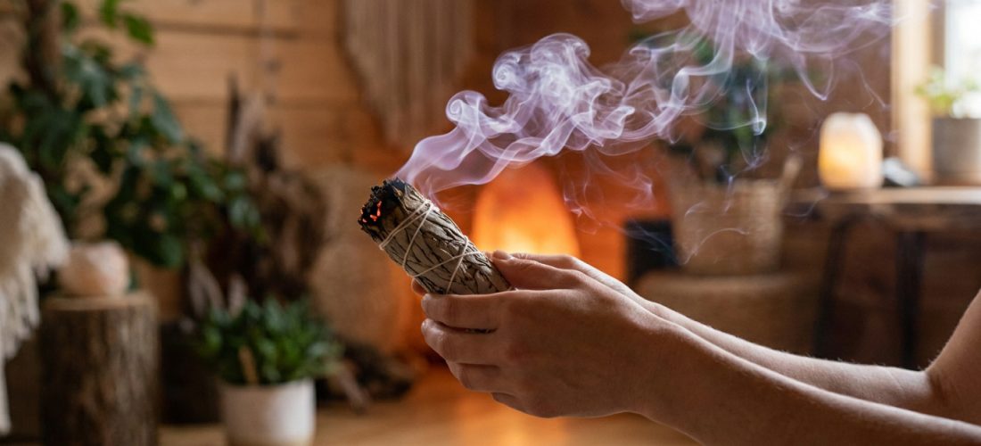 Des mains tiennent un bâton de sauge blanche allumé, d'où s'échappe une fumée violette ondulante. Ambiance intérieure boisée et chaleureuse.