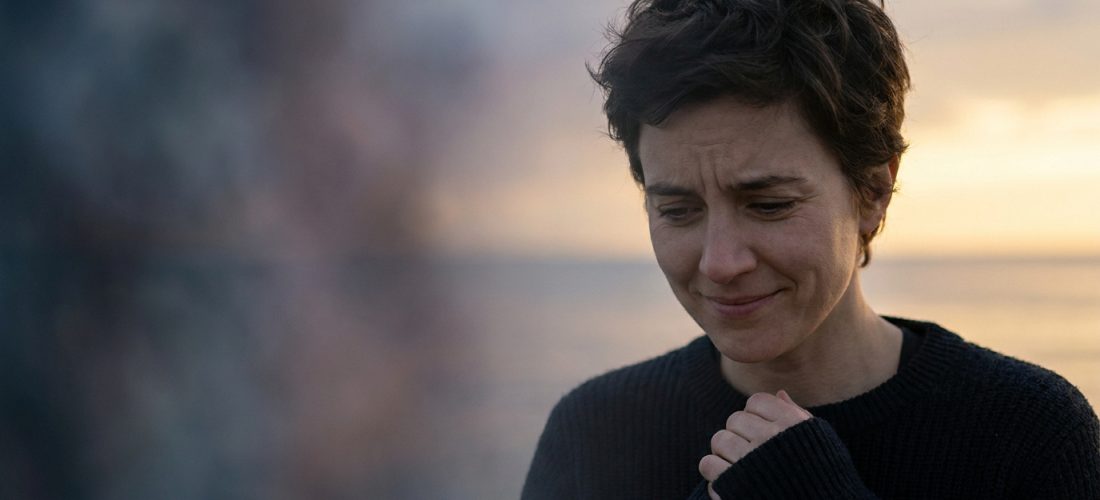 Portrait d'une femme aux cheveux courts, les mains jointes et le regard baissé, semblant pensive ou inquiète devant un coucher de soleil flou.